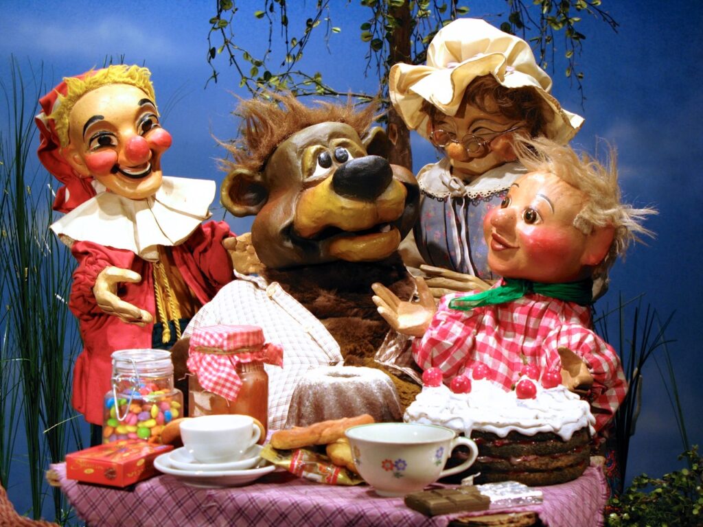 Kasperl, Bernie der Bär, Großmutti und Sepperl vor einem reich gedeckten Tisch mit Geburtstagstorte und anderen Leckereien
